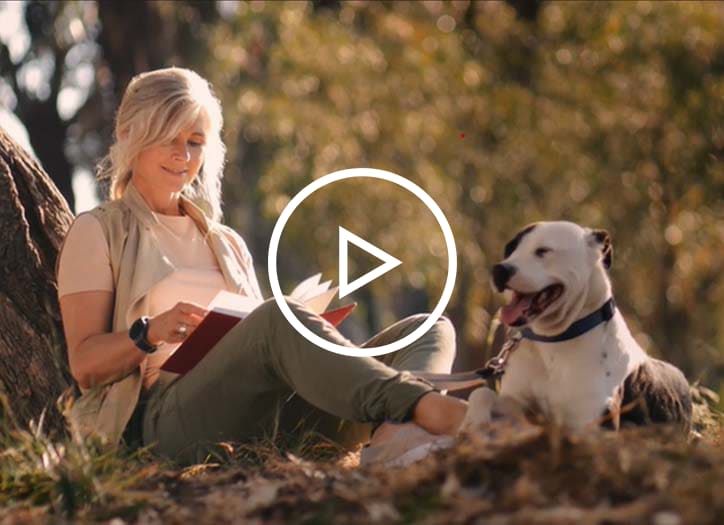 Woman sits under tree and reads as her dog lies next to her