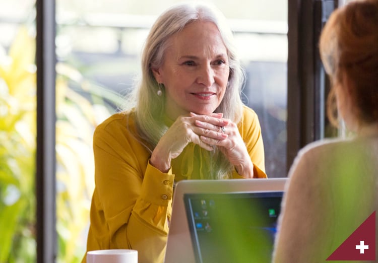 Senior business woman working on laptop smiles at coworker across a desk