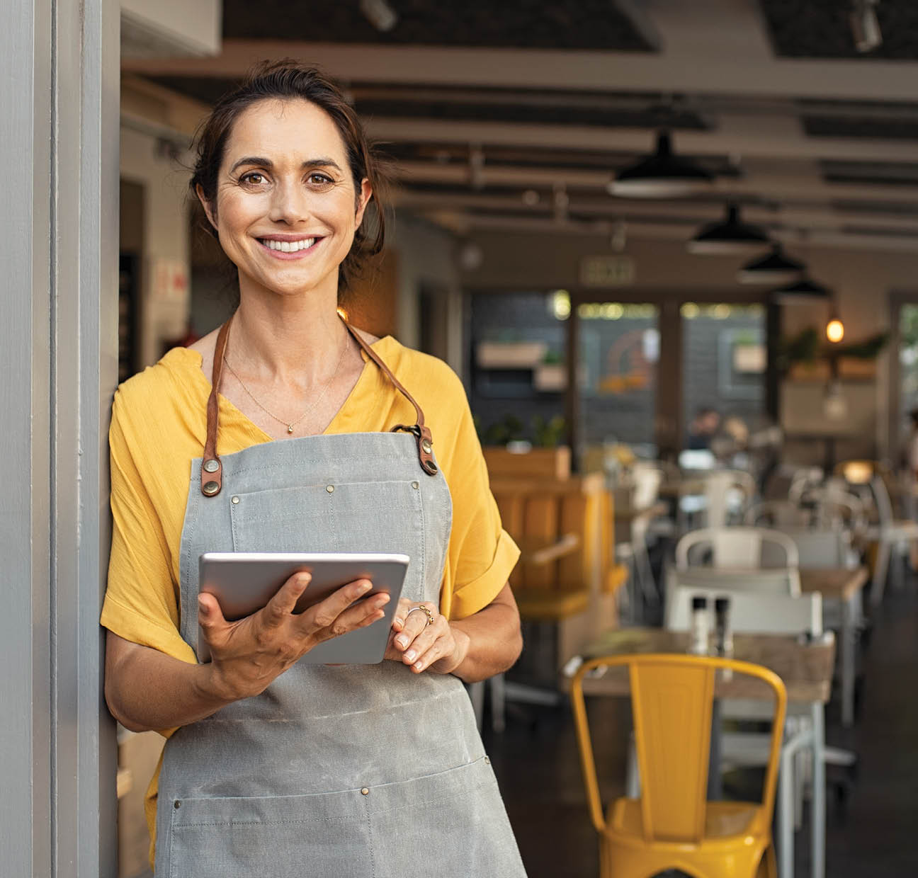 Portrait of happy woman standing at doorway of her store holding digital tablet  Cheerful mature waitress waiting for clients at coffee shop  Successful small business owner in casual clothing and grey apron standing at entrance and looking at camera 