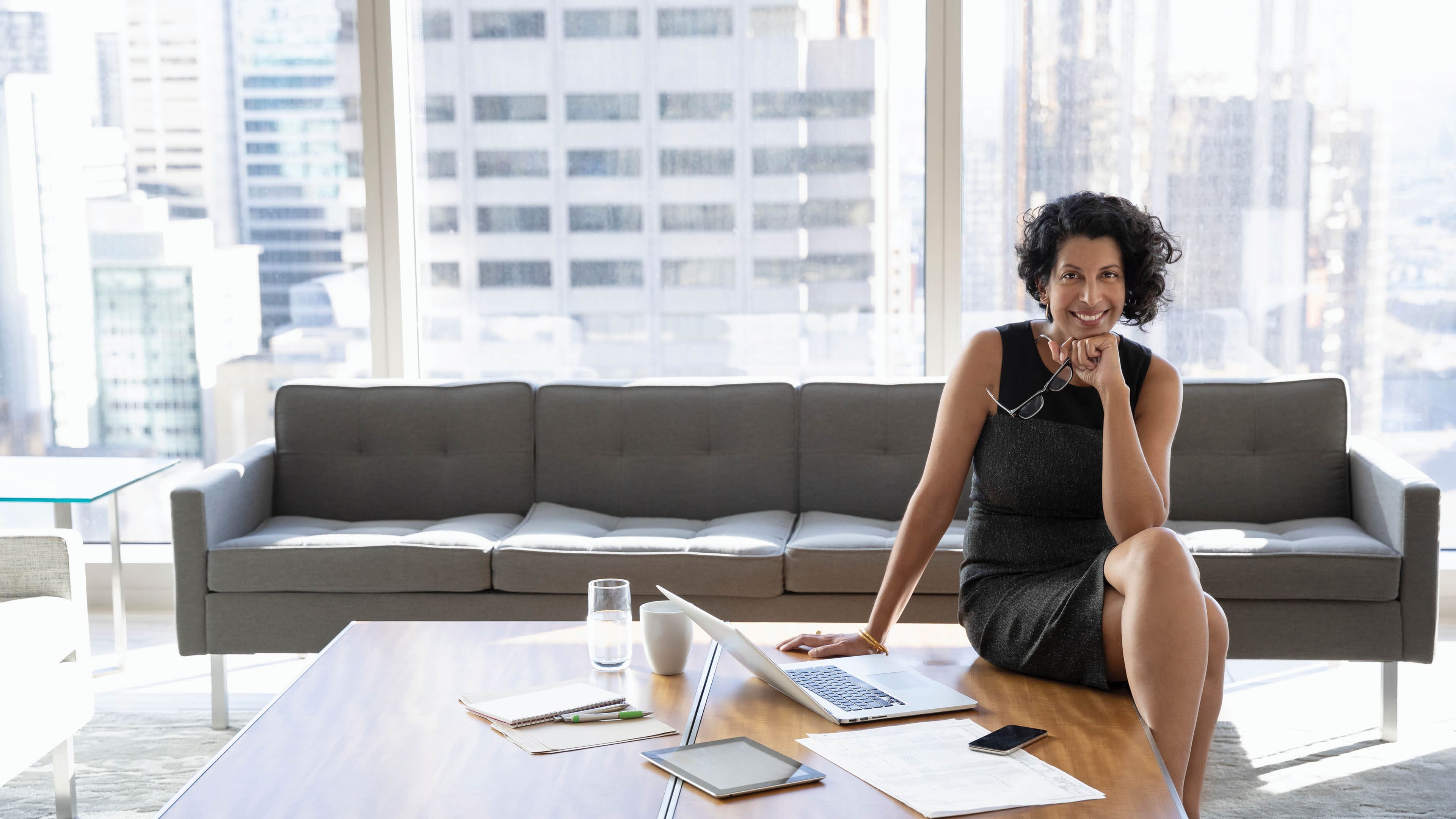 A smiling businesswoman is sitting on a desk next to a laptop, in a modern high rise office.