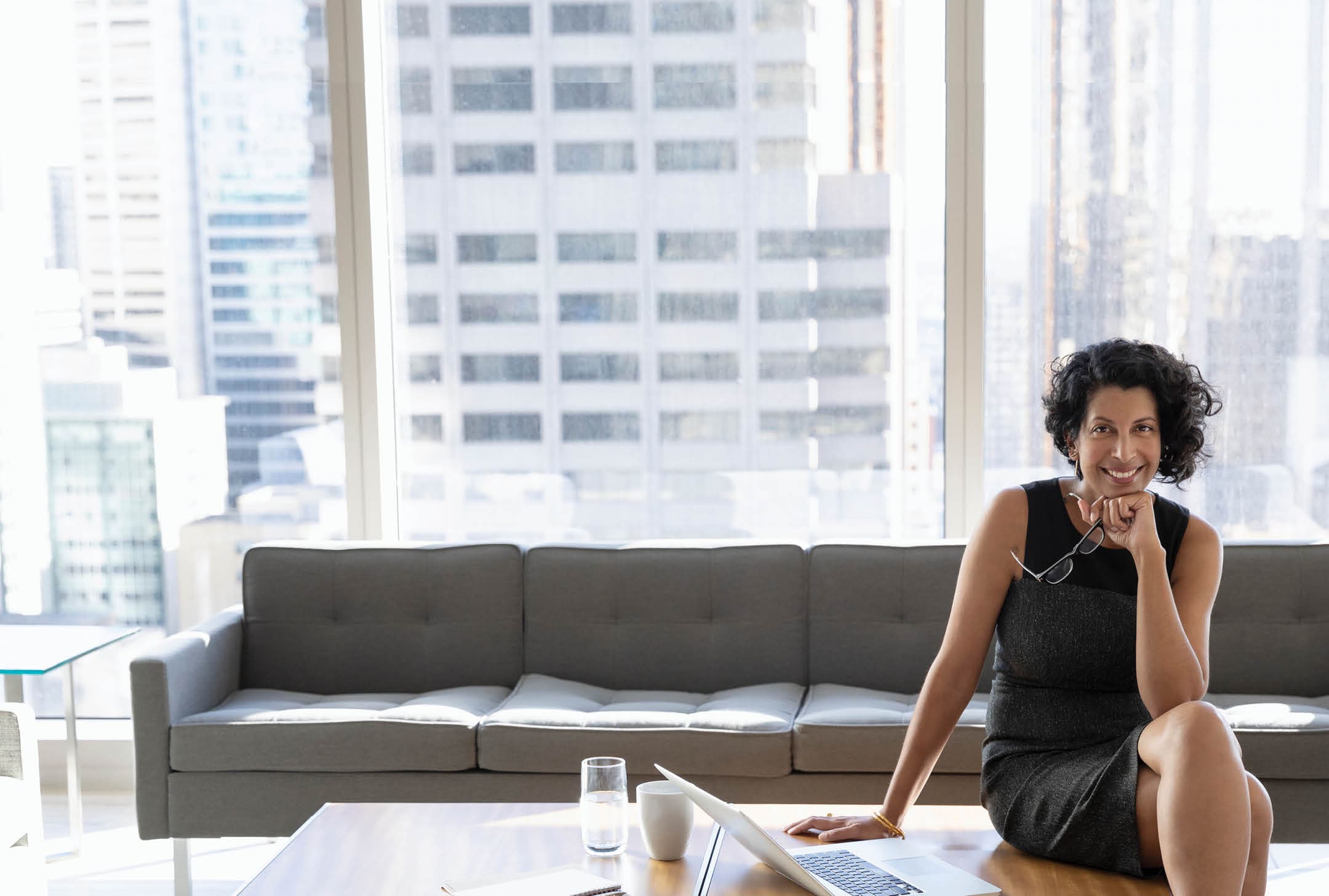 A smiling businesswoman is sitting on a desk next to a laptop, in a modern high rise office.