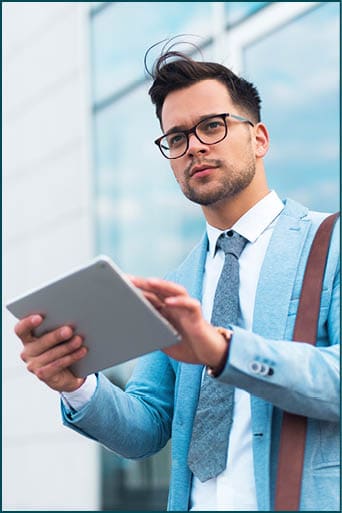 Serious businessman in suit standing on the street looking around while holding tablet in his hands.