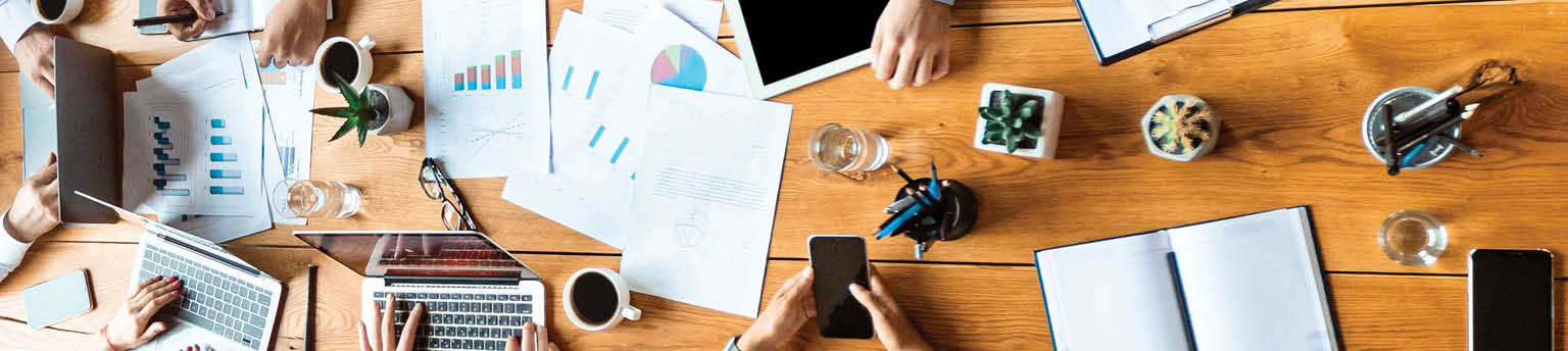 Image of office table filled with pens, computers, paperwork and hands. 