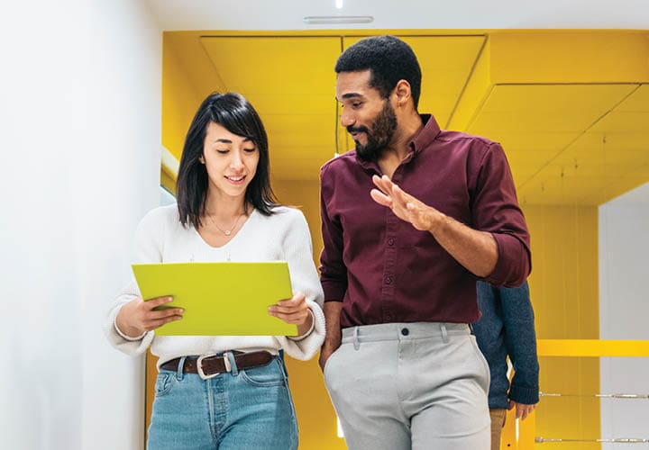 two colleagues looking at a printin out while walking down an office hall